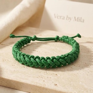 May include: A dark green braided cord bracelet featuring a woven pattern and adjustable tie ends. The bracelet is positioned near a card with the text Vera by Mila printed in a simple font, showcasing the detailed texture of the green cord.