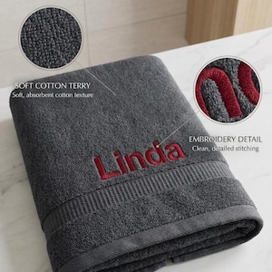 May include: A dark gray cotton terry towel featuring the embroidered name Linda in red thread. The towel has a textured surface and a horizontal ribbed border detail. Two circular insets highlight the soft cotton texture and the embroidery detail.