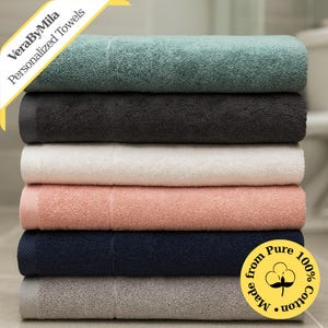 May include: A stack of six plush bath towels in various colors: teal, black, white, peach, navy, and gray. The towels are made of 100% cotton and feature a textured design. The text "VeraByMila Personalized Towels" is visible.