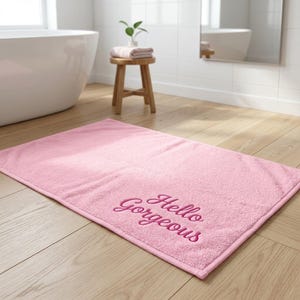 May include: A rectangular light pink bath mat with a textured fabric surface. The words "Hello Gorgeous" are embroidered in a darker pink script font in the bottom right corner of the mat, which is placed on a light wood floor.