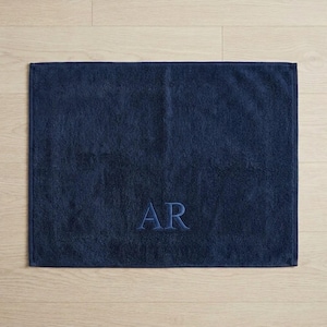 May include: A rectangular navy blue bath towel featuring a textured terry cloth surface. The center of the towel is embroidered with the capital letters AR in a matching navy blue thread, creating a subtle and uniform appearance.