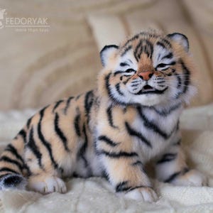 May include: A plush tiger cub with a smiling expression, featuring a tan and black striped pattern. The toy is sitting on a white, textured surface. The text "ELENA FEDORYAK more than toys" is visible in the background.