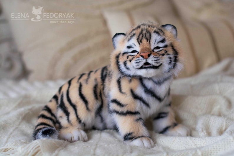 May include: A plush tiger cub with a smiling expression, featuring a tan and black striped pattern. The toy is sitting on a white, textured surface. The text "ELENA FEDORYAK more than toys" is visible in the background.