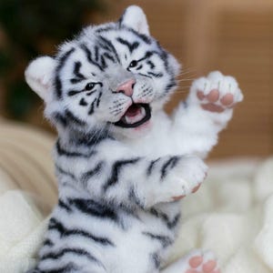 Könnte beinhalten: Ein weißes Tigerbaby aus Plüsch mit schwarzen Streifen, offenem Mund und erhobener Pfote. Das Junge hat eine rosa Nase und Pfotenballen. Das Bild enthält den Text "ELENA FEDORYAK more than toys".