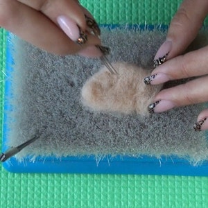 Puede incluir: Se muestran las manos de una persona felpando un peque&ntilde;o trozo de lana marr&oacute;n claro con una aguja de felpado sobre una alfombra de felpado gris. La alfombra est&aacute; sobre una superficie verde.