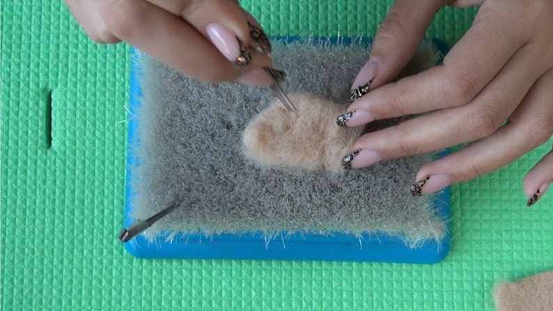 Puede incluir: Se muestran las manos de una persona felpando un peque&ntilde;o trozo de lana marr&oacute;n claro con una aguja de felpado sobre una alfombra de felpado gris. La alfombra est&aacute; sobre una superficie verde.
