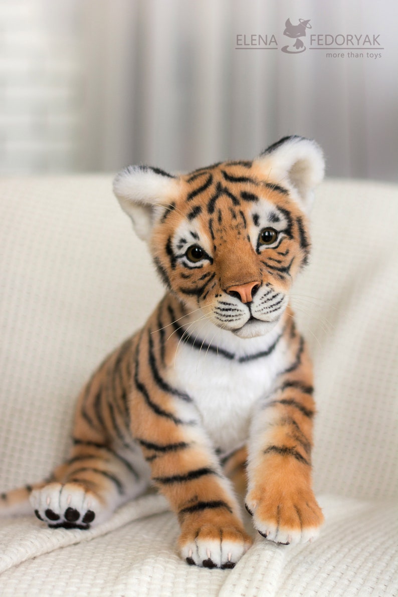 May include: A plush tiger cub toy with orange and black stripes, white fur on its chest and belly, and black paw pads. The toy is sitting on a white fluffy surface.