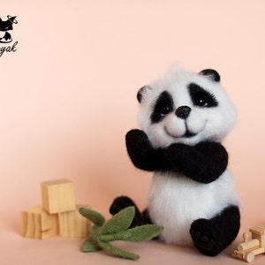May include: A small, white and black panda bear plush toy with a smiling face. The panda is sitting on a pink surface with wooden blocks and green leaves.