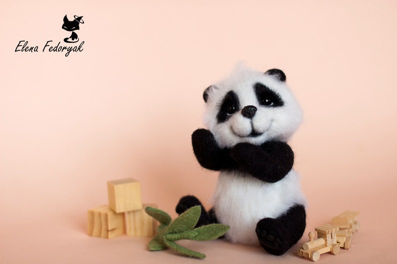 May include: A small, white and black panda bear plush toy with a smiling face. The panda is sitting on a pink surface with wooden blocks and green leaves.