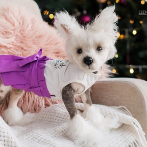 May include: A small white dog wearing a white tank top with a purple skirt. The dog is sitting on a white blanket in front of a Christmas tree.