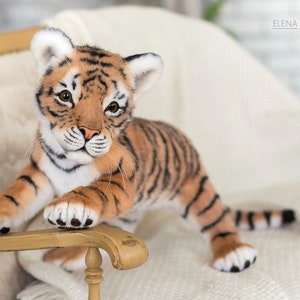 May include: A plush tiger cub with orange and black stripes sits on a white chair. The tiger cub has a white chest and paws and is looking directly at the camera.