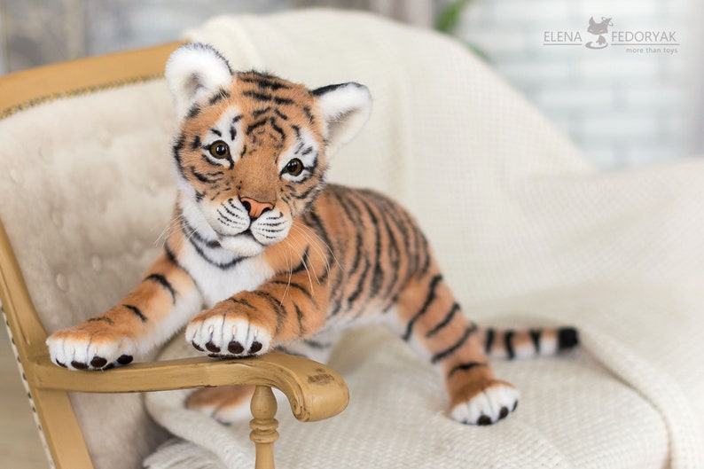 May include: A plush tiger cub with orange and black stripes sits on a white chair. The tiger cub has a white chest and paws and is looking directly at the camera.