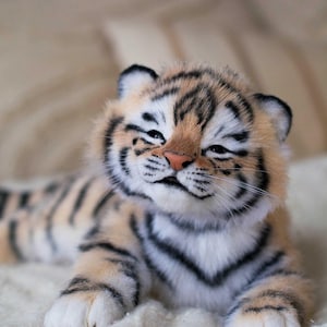 May include: A plush tiger cub with a light orange and black striped pattern. The cub has a pink nose, white paws, and is lying on a white surface. The text "ELENA FEDORYAK more than toys" is visible.
