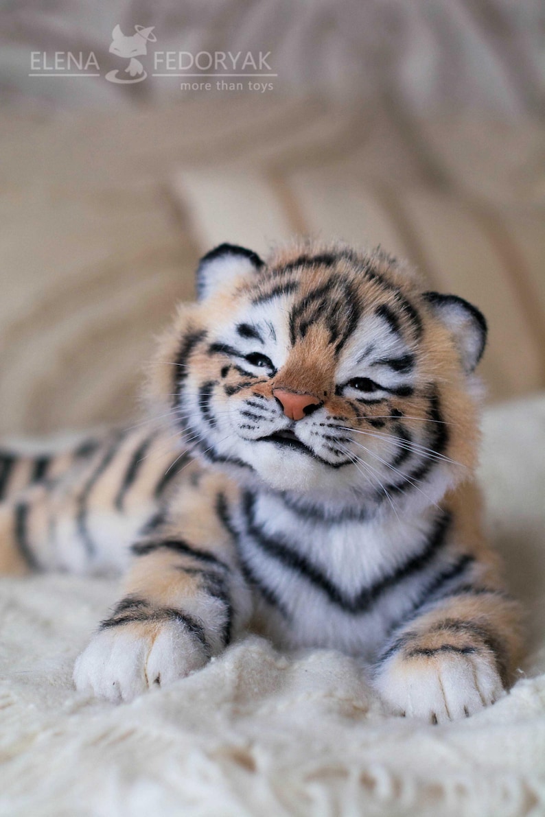 May include: A plush tiger cub with a light orange and black striped pattern. The cub has a pink nose, white paws, and is lying on a white surface. The text "ELENA FEDORYAK more than toys" is visible.