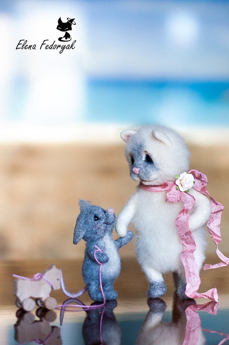May include: A white and gray cat with blue eyes and a pink ribbon is standing next to a gray mouse with a pink string. The mouse is pulling a wooden toy horse.