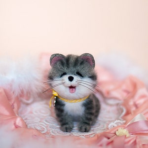 May include: A gray and white needle felted cat with a yellow collar and a pink tongue sticking out. The cat is sitting on a pink and white fluffy surface.