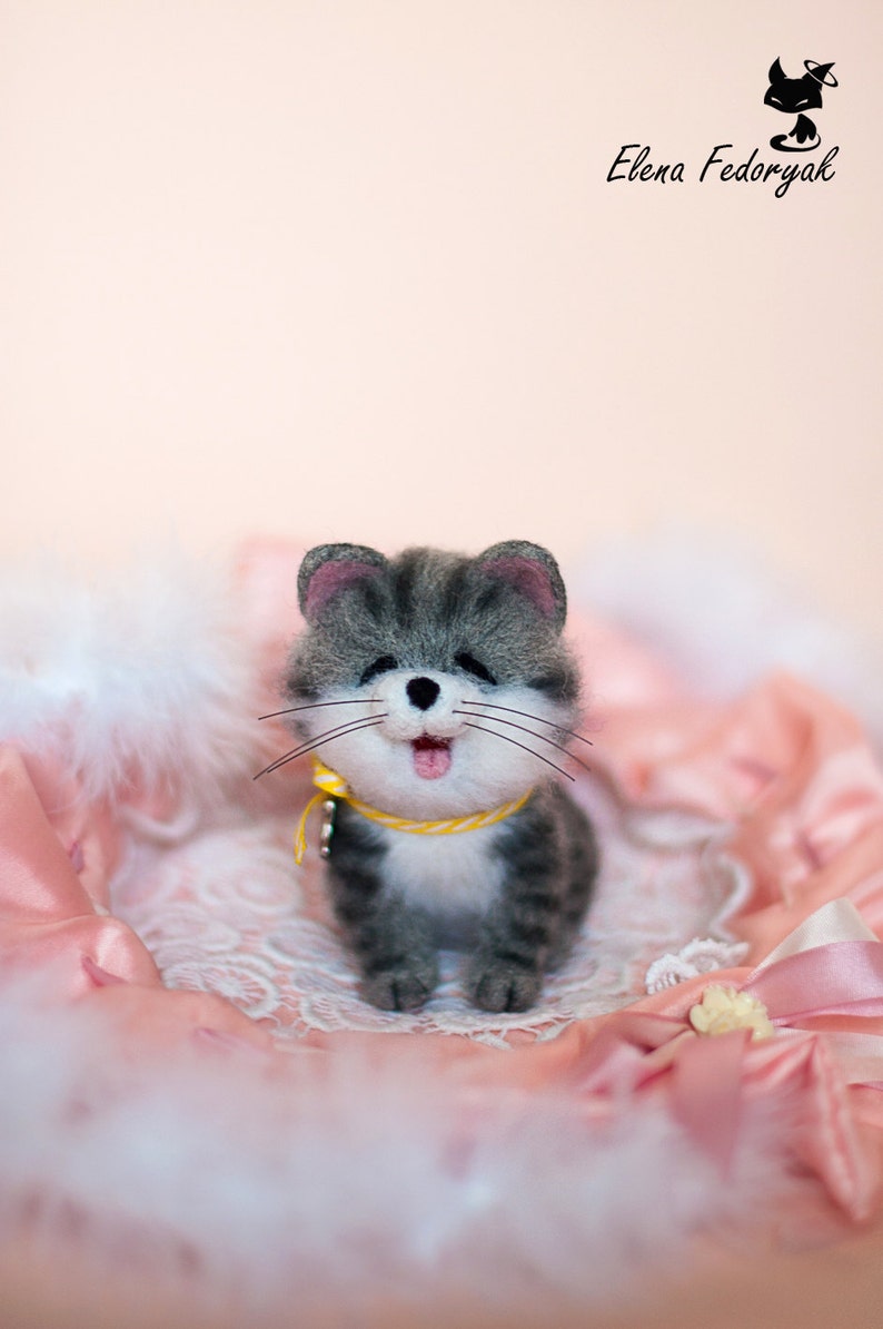 May include: A gray and white needle felted cat with a yellow collar and a pink tongue sticking out. The cat is sitting on a pink and white fluffy surface.