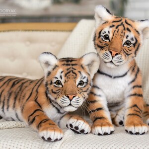 May include: Two realistic plush tiger cubs, one lying down and the other sitting up, both looking at the camera. They are both orange and black with white stripes and have black noses and paws.
