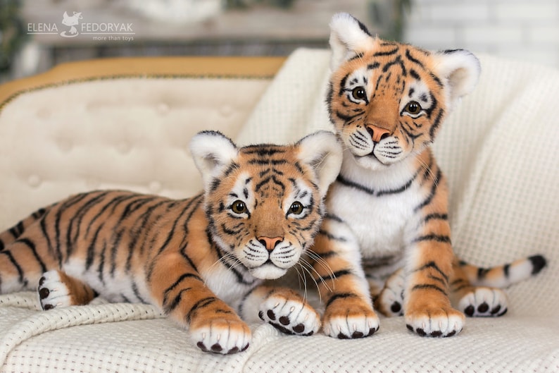 May include: Two realistic plush tiger cubs, one lying down and the other sitting up, both looking at the camera. They are both orange and black with white stripes and have black noses and paws.