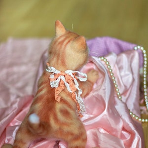 Puede incluir: Un gato atigrado jengibre con un lazo blanco alrededor del cuello est&aacute; sentado sobre una tela de sat&eacute;n rosa. El gato est&aacute; mirando hacia arriba y hacia la derecha.