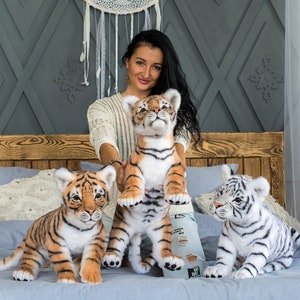 May include: Three plush tiger cubs, one white and two orange and black, are sitting on a bed. A woman is holding the larger orange and black tiger cub.