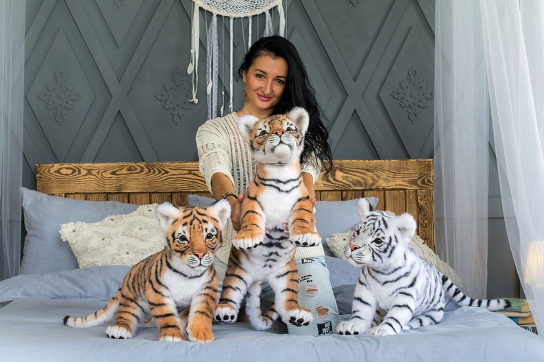 May include: Three plush tiger cubs, one white and two orange and black, are sitting on a bed. A woman is holding the larger orange and black tiger cub.