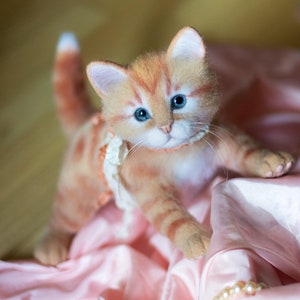 May include: A close-up of a ginger and white felted cat wearing a pink and white lace collar. The cat is sitting on a pink satin fabric with a pearl necklace.