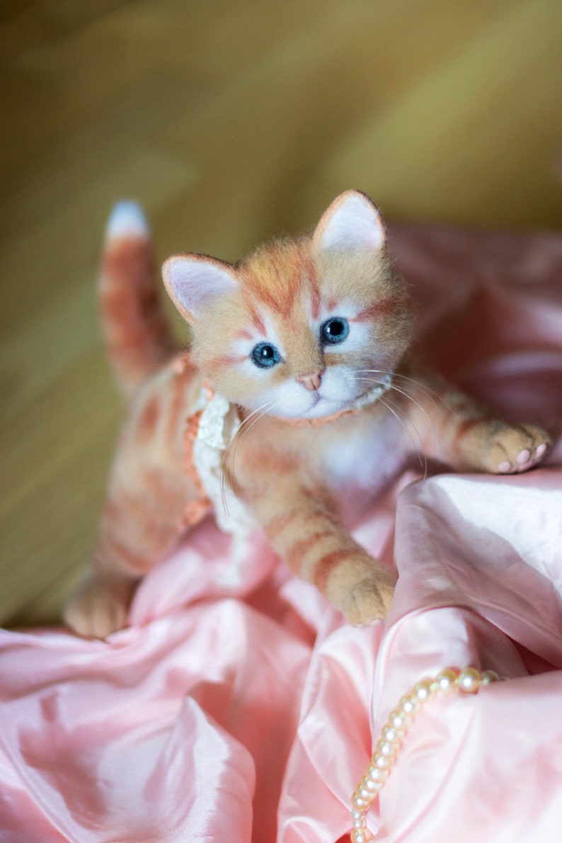 May include: A close-up of a ginger and white felted cat wearing a pink and white lace collar. The cat is sitting on a pink satin fabric with a pearl necklace.