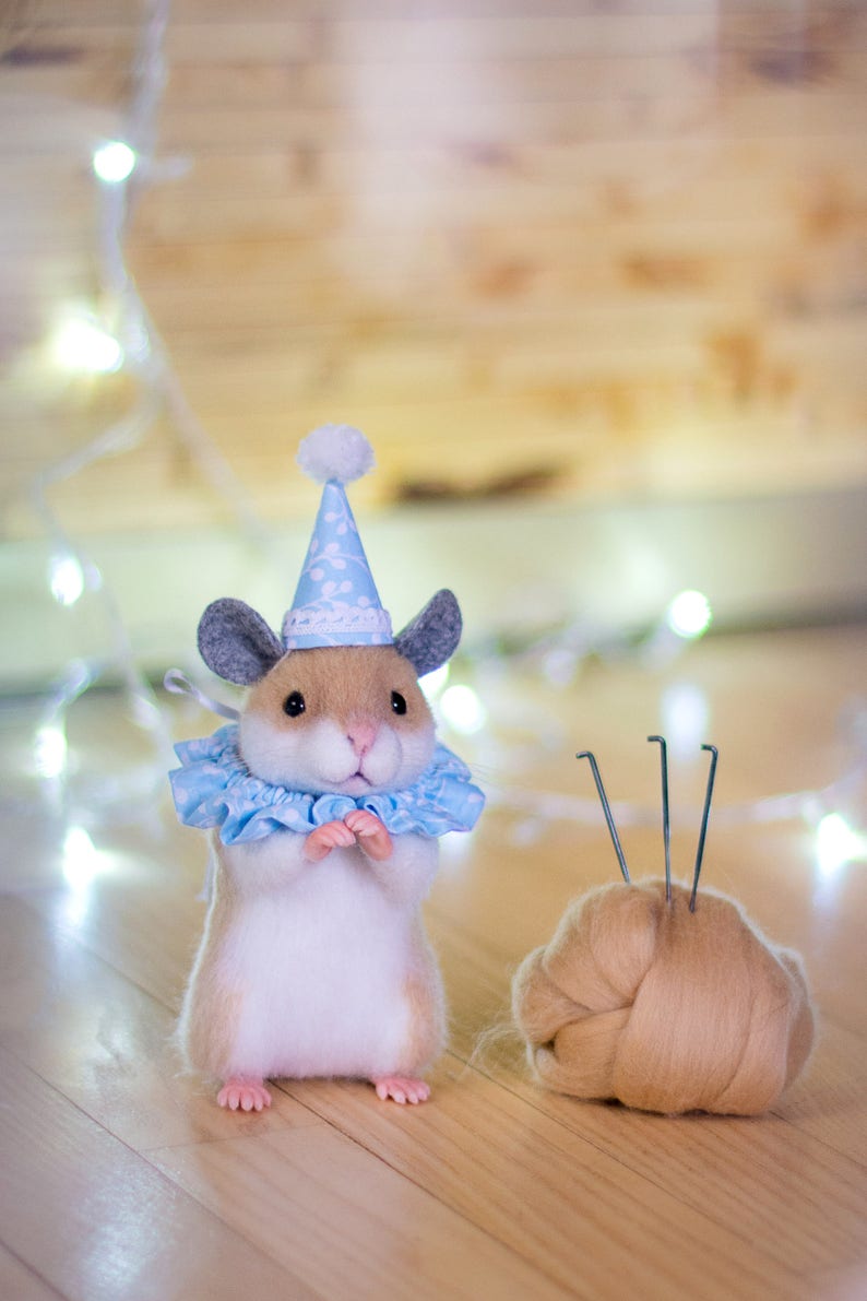 May include: A small, white and brown hamster wearing a blue and white party hat and a blue and white ruffled collar. The hamster is standing on a wooden surface with a ball of brown wool and needles in the background.