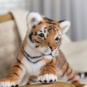 May include: A realistic plush tiger cub with orange and black stripes, white fur on its chest and face, and black paw pads. The tiger cub is sitting on a wooden chair with a white blanket.