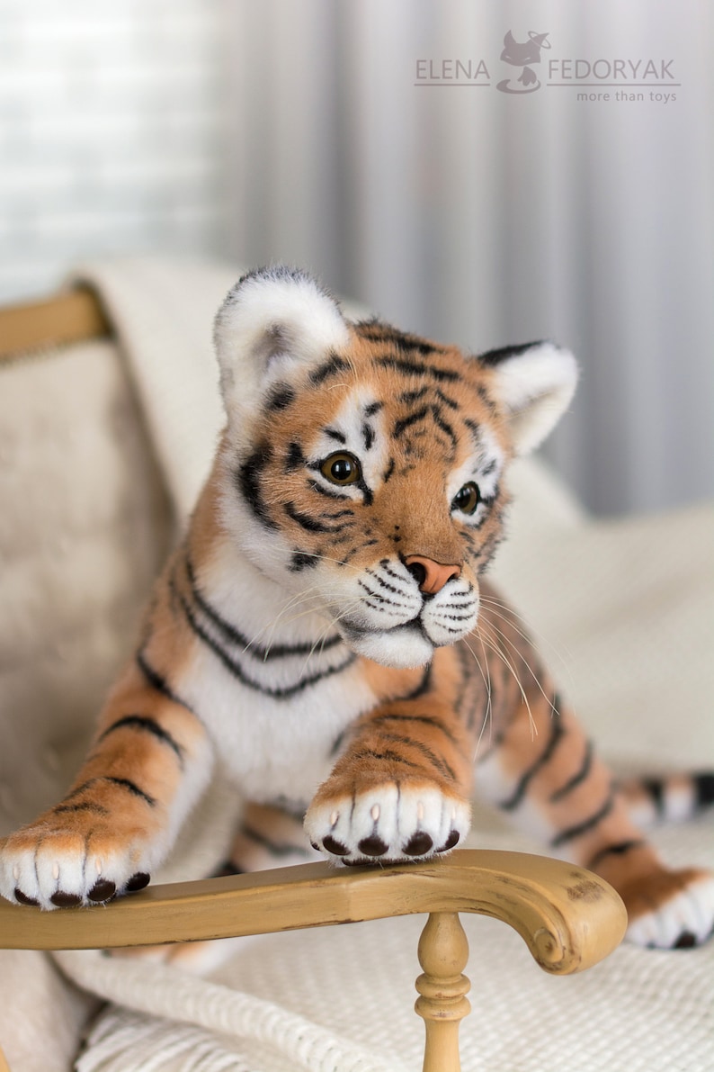 May include: A realistic plush tiger cub with orange and black stripes, white fur on its chest and face, and black paw pads. The tiger cub is sitting on a wooden chair with a white blanket.