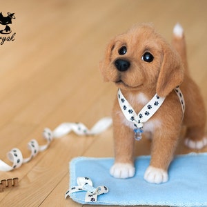 May include: A light brown felt puppy wearing a white collar with black paw prints and a blue charm. The puppy is sitting on a blue felt mat with a white ribbon tied in a bow. The puppy is looking to the right of the image. The word "Малыш" is written in brown letters on a wooden surface.