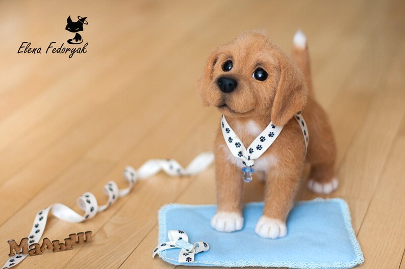May include: A light brown felt puppy wearing a white collar with black paw prints and a blue charm. The puppy is sitting on a blue felt mat with a white ribbon tied in a bow. The puppy is looking to the right of the image. The word "Малыш" is written in brown letters on a wooden surface.