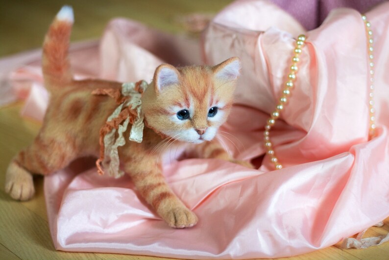 May include: A ginger tabby kitten wearing a lace collar and a bow, sitting on a pink satin fabric with a pearl necklace.