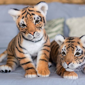 May include: Two realistic plush tiger cubs, one sitting and one lying down, with orange and black stripes and white accents. They are on a light blue fabric surface.