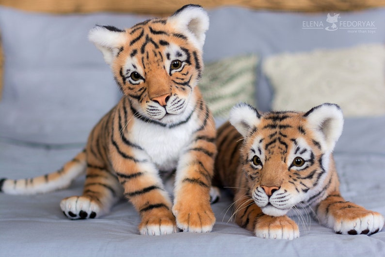 May include: Two realistic plush tiger cubs, one sitting and one lying down, with orange and black stripes and white accents. They are on a light blue fabric surface.
