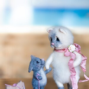 Puede incluir: Un gato blanco y gris con ojos azules y una cinta rosa est&aacute; de pie junto a un rat&oacute;n gris con una cuerda rosa. El rat&oacute;n est&aacute; tirando de un caballo de juguete de madera.