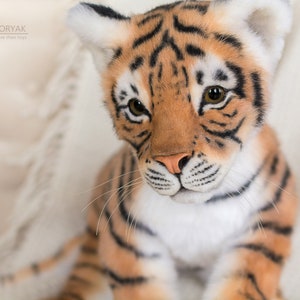 May include: Close-up of a plush tiger cub with orange and black stripes. The tiger has a white chest and a pink nose. The tiger is looking at the camera.