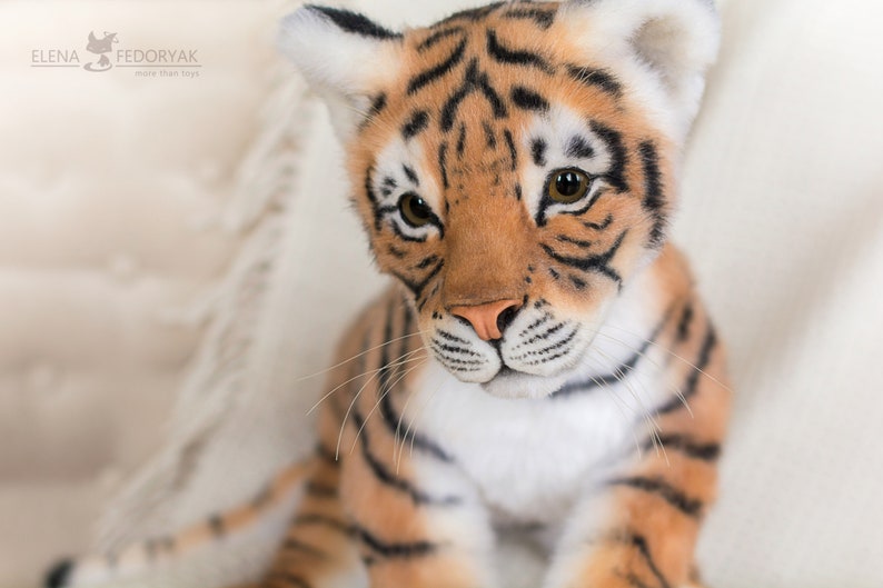 May include: Close-up of a plush tiger cub with orange and black stripes. The tiger has a white chest and a pink nose. The tiger is looking at the camera.