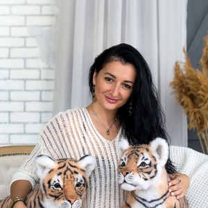 May include: A woman is sitting on a sofa with two plush tiger cubs. The tiger cubs are brown and orange with black stripes. The woman is wearing a white sweater and blue jeans. The tiger cubs are looking at the camera.
