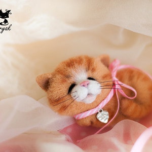 May include: A small, orange and white needle felted cat wearing a pink ribbon with a silver heart charm. The cat is lying on a pink and white fabric.
