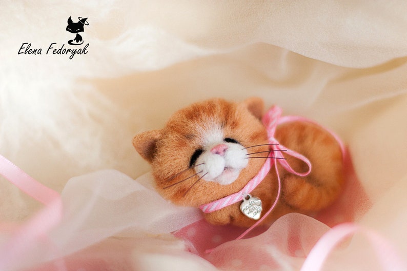 May include: A small, orange and white needle felted cat wearing a pink ribbon with a silver heart charm. The cat is lying on a pink and white fabric.