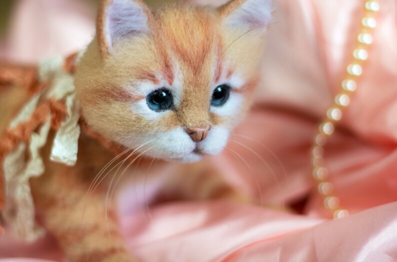 Puede incluir: Primer plano de un gatito jengibre con ojos azules que lleva un lazo de color melocot&oacute;n. El gatito est&aacute; tumbado sobre una tela de sat&eacute;n rosa.