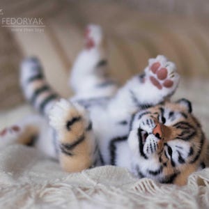 May include: A plush tiger cub toy lying on its back with its paws in the air. The toy has a white and orange striped pattern, with a pink nose and paw pads. The toy is lying on a cream-colored blanket.