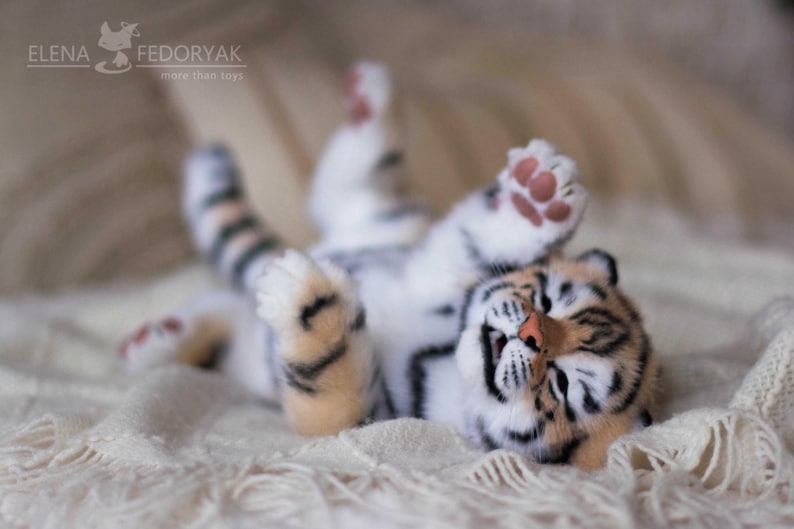 May include: A plush tiger cub toy lying on its back with its paws in the air. The toy has a white and orange striped pattern, with a pink nose and paw pads. The toy is lying on a cream-colored blanket.