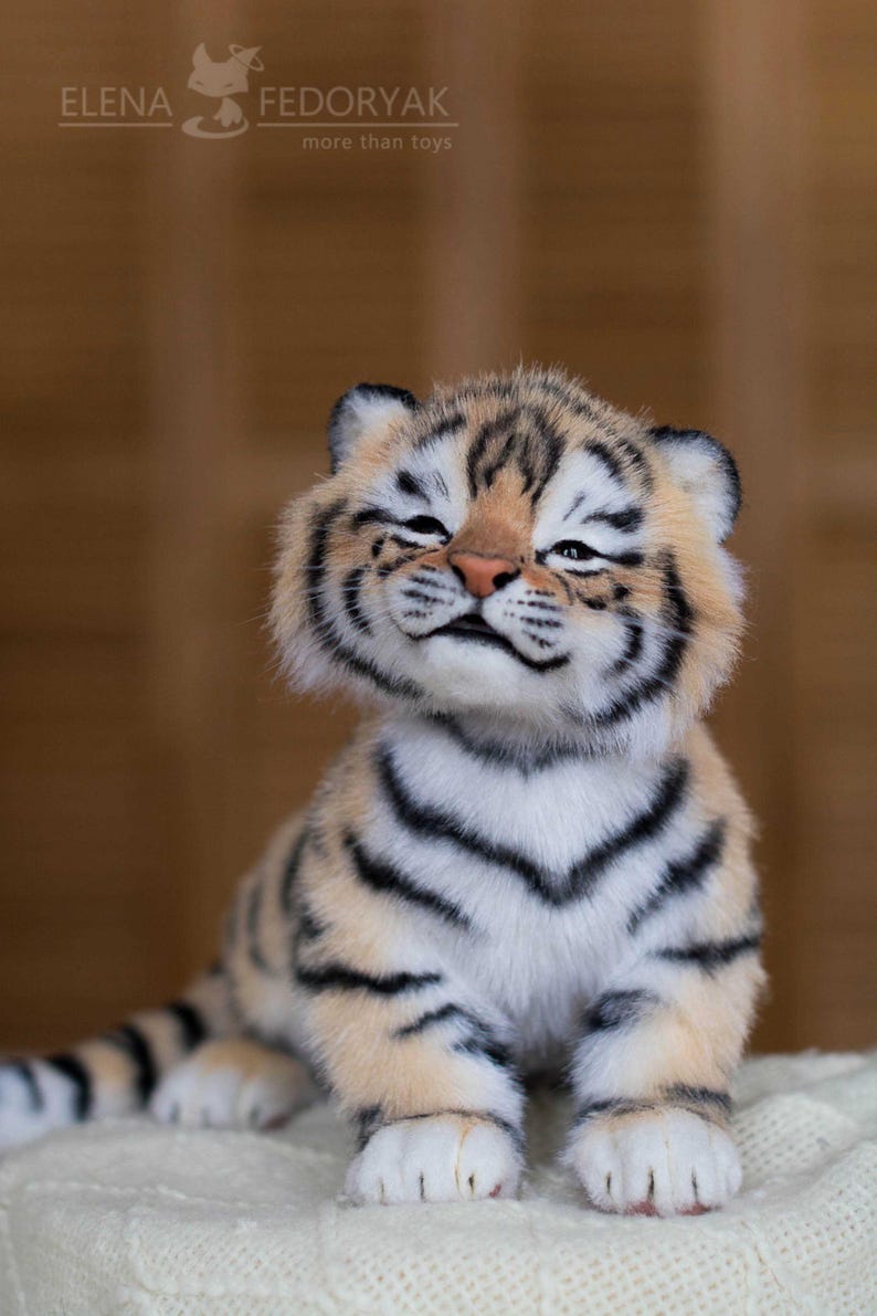 May include: A plush tiger cub with a smiling expression. The cub has a beige and white body with black stripes. The text "ELENA FEDORYAK more than toys" is visible in the upper left corner.