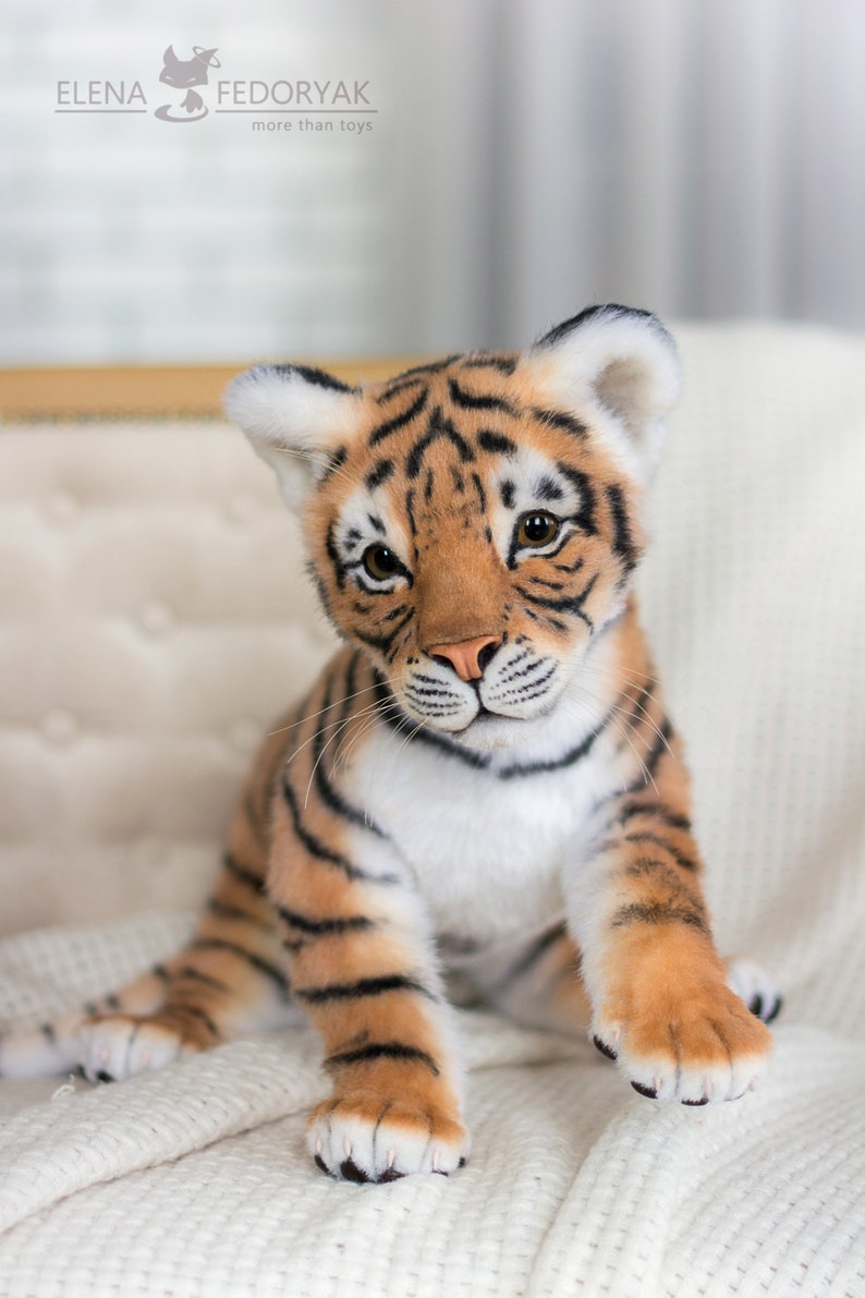 May include: A plush tiger cub toy with orange and black stripes, white fur on its chest and belly, and a sweet expression. The toy is sitting on a white fluffy surface.