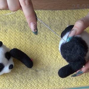 Puede incluir: Una persona est&aacute; haciendo fieltro con aguja un oso panda blanco y negro. El panda se est&aacute; haciendo fieltro en una alfombra amarilla. La persona est&aacute; usando una herramienta de fieltro con aguja para dar forma al pelaje del panda.