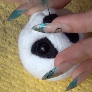 Puede incluir: Primer plano de una cabeza de panda de fieltro blanco con ojos y nariz de fieltro negro. La cabeza de panda est&aacute; siendo sostenida por una mano con u&ntilde;as largas y decoradas.