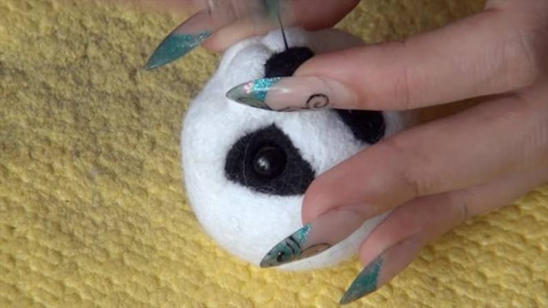 Puede incluir: Primer plano de una cabeza de panda de fieltro blanco con ojos y nariz de fieltro negro. La cabeza de panda est&aacute; siendo sostenida por una mano con u&ntilde;as largas y decoradas.